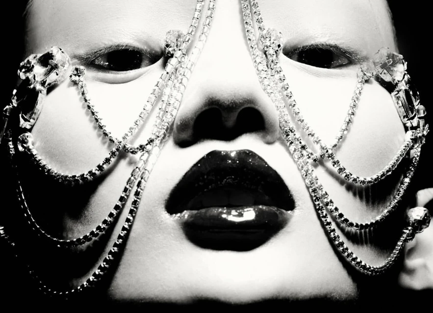 Close-up portrait of a face with a jeweled face covering, glossy lips, pale skin, and dark eyes, shot in black and white.