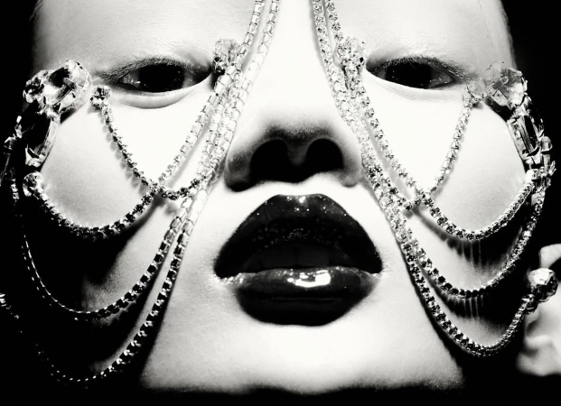 Close-up portrait of a face with a jeweled face covering, glossy lips, pale skin, and dark eyes, shot in black and white.