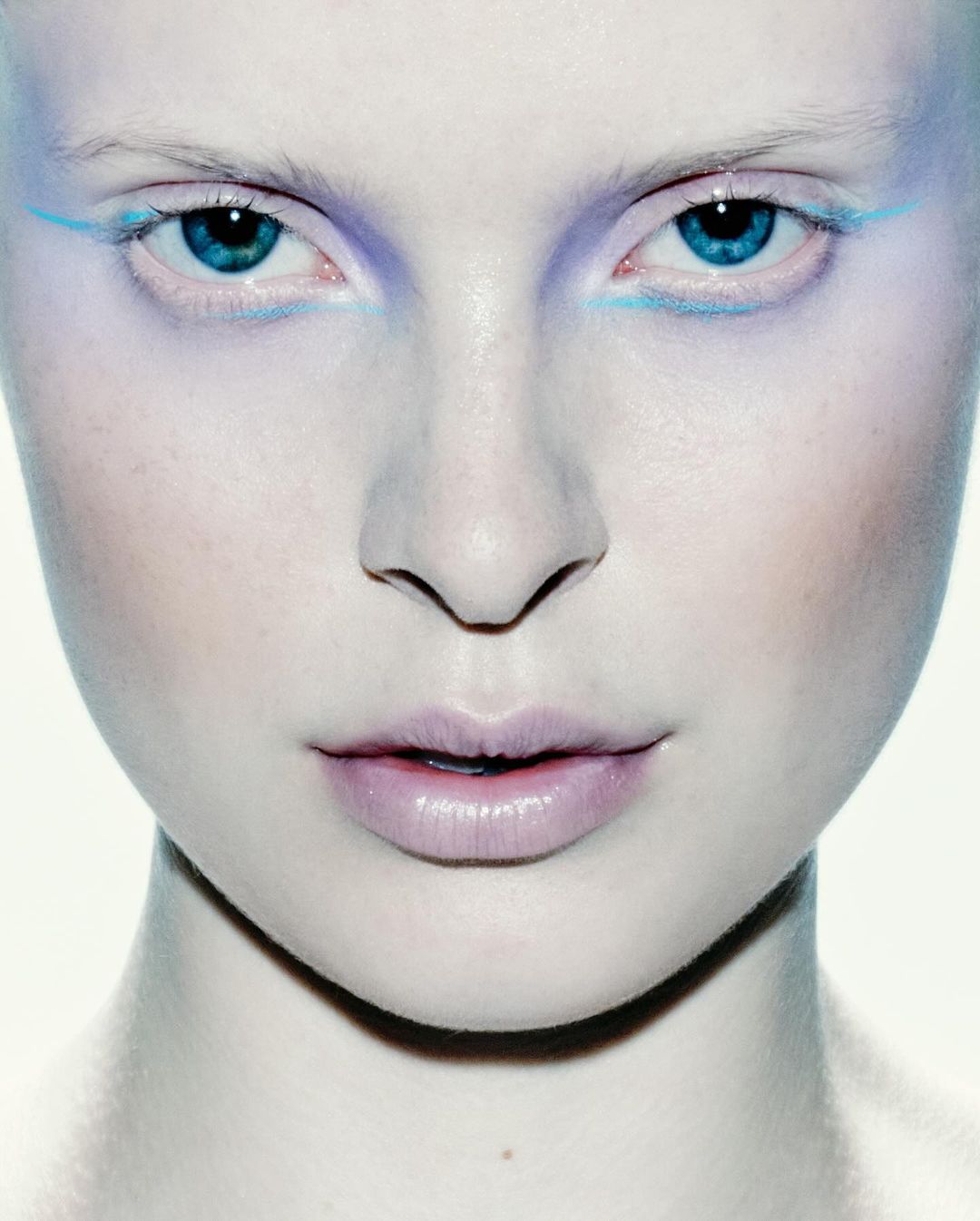 Close-up of a woman's face with light skin and blue eyes, accented with blue and purple makeup, against a white background.