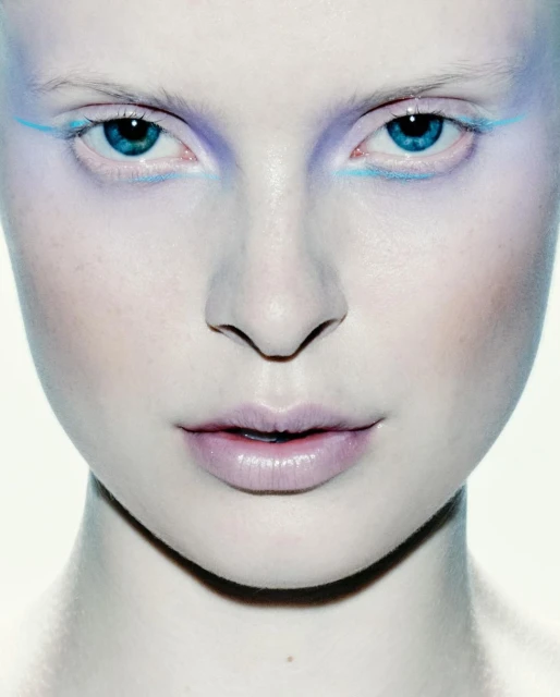 Close-up of a woman's face with light skin and blue eyes, accented with blue and purple makeup, against a white background.