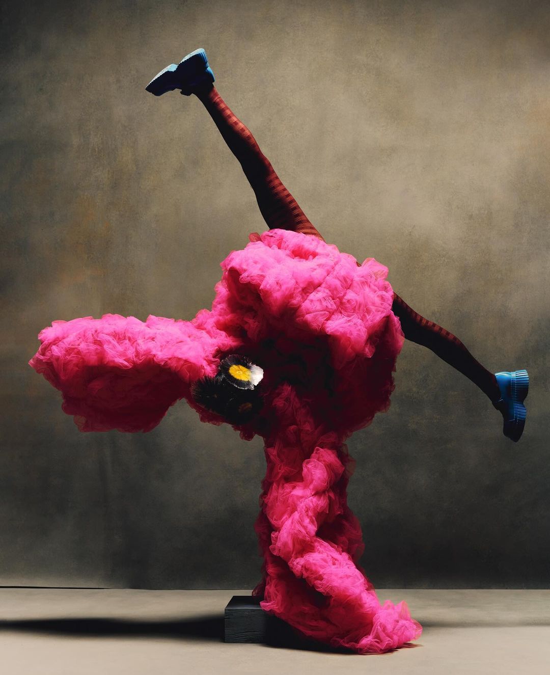 Abstract fashion image of a person in an unusual pink ruffle costume wearing blue shoes, balancing on a black stand in studio lighting.