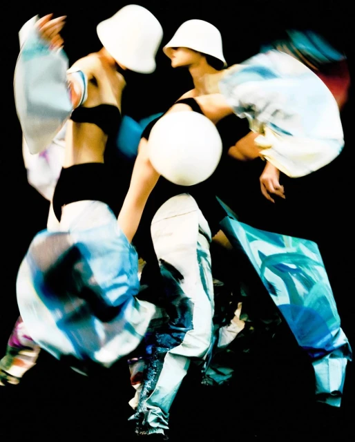 Two models in motion wearing hats and blue and white patterned outfits against a black backdrop, capturing dynamic fashion photography.