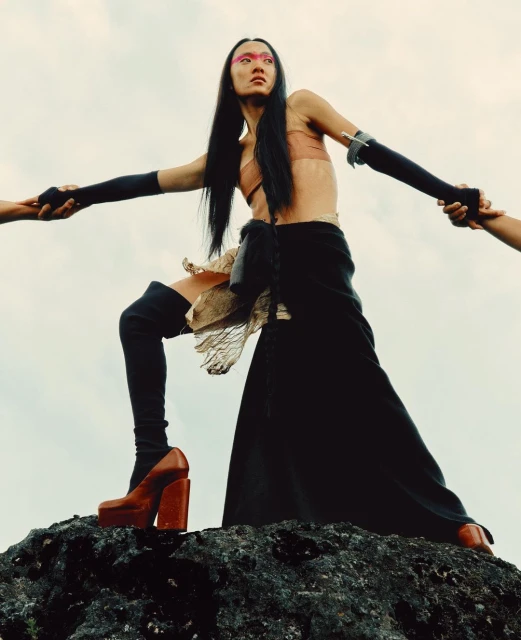 A striking fashion shot shows a model in avant-garde apparel on a rock against a cloudy sky, emphasizing unique design and texture.