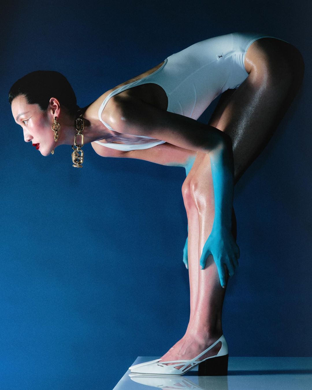 Woman in white swimsuit poses in a blue studio with blue skin paint, statement earrings, and matching white heels, creating a surreal fashion shot.