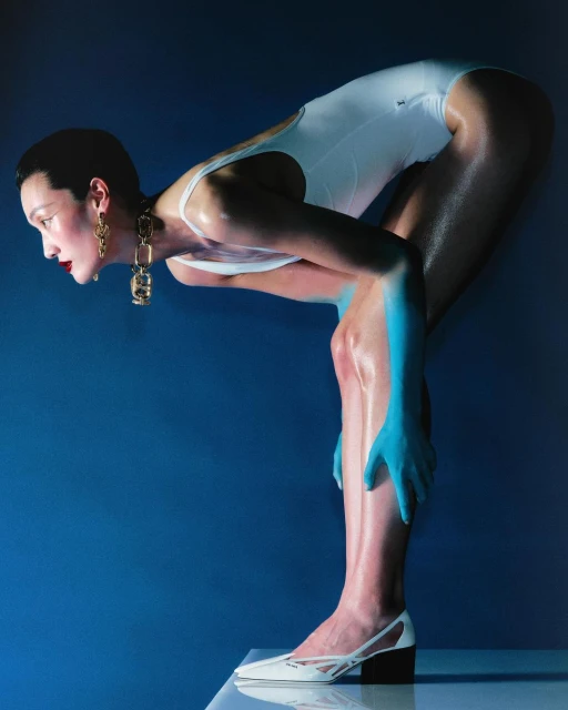 Woman in white swimsuit poses in a blue studio with blue skin paint, statement earrings, and matching white heels, creating a surreal fashion shot.