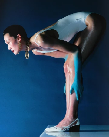 Woman in white swimsuit poses in a blue studio with blue skin paint, statement earrings, and matching white heels, creating a surreal fashion shot.