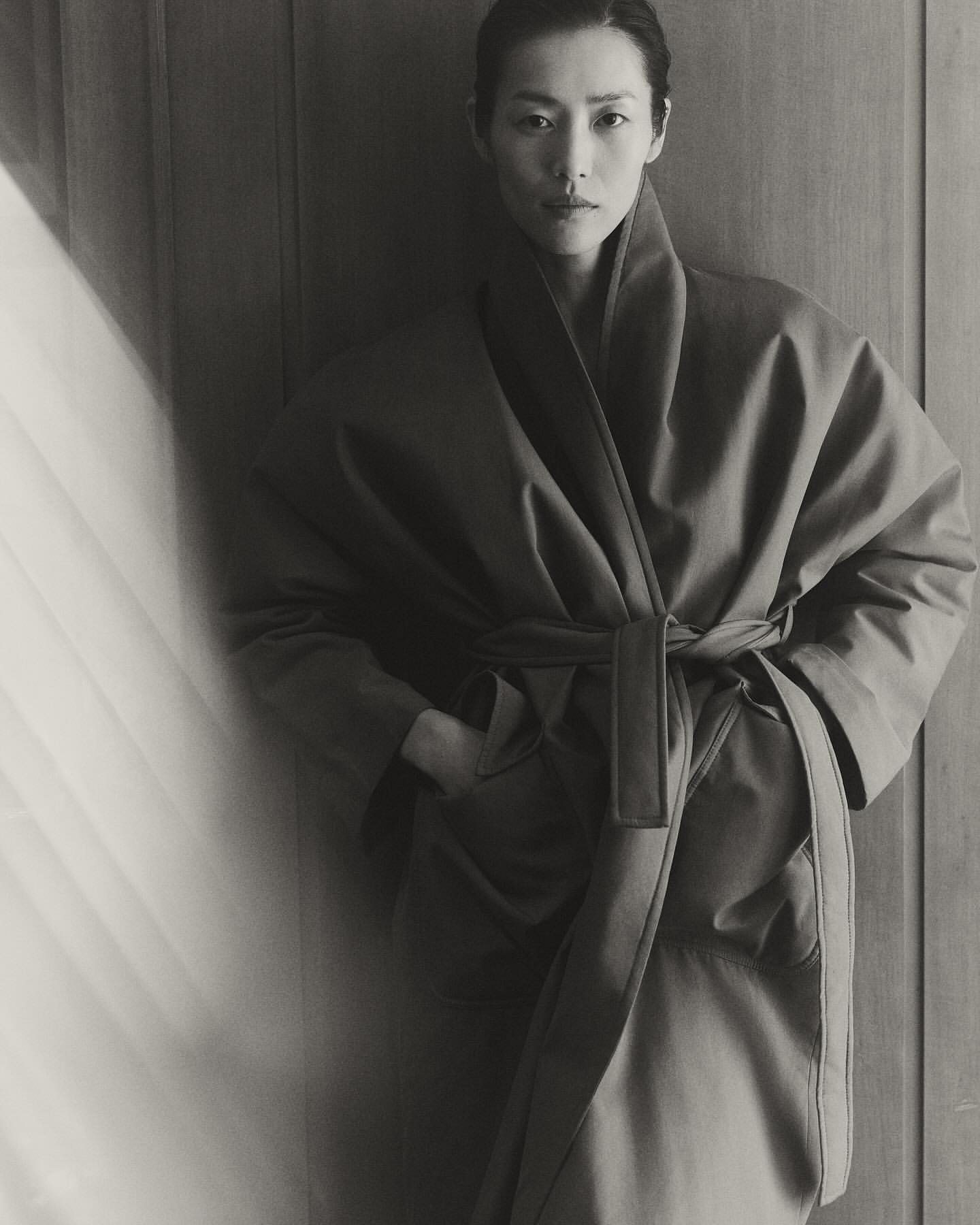 Monochrome portrait of an Asian woman in a robe with a sash, hands in pockets, standing indoors with soft, natural light and wood background.