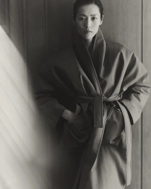 Monochrome portrait of an Asian woman in a robe with a sash, hands in pockets, standing indoors with soft, natural light and wood background.