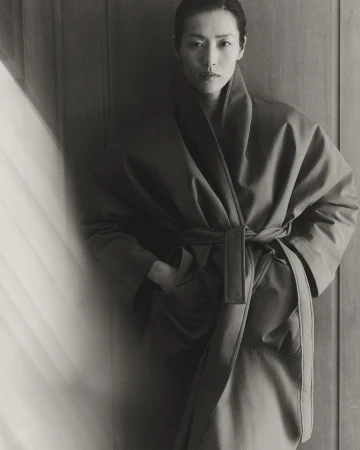 Monochrome portrait of an Asian woman in a robe with a sash, hands in pockets, standing indoors with soft, natural light and wood background.