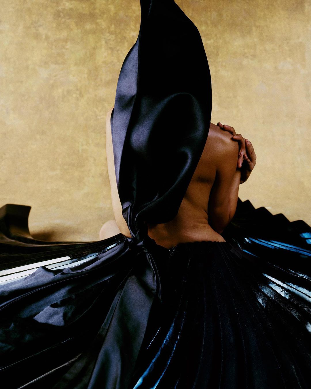 Back view of a partially visible Black woman wearing a draped black dress against a golden background in a studio setting.