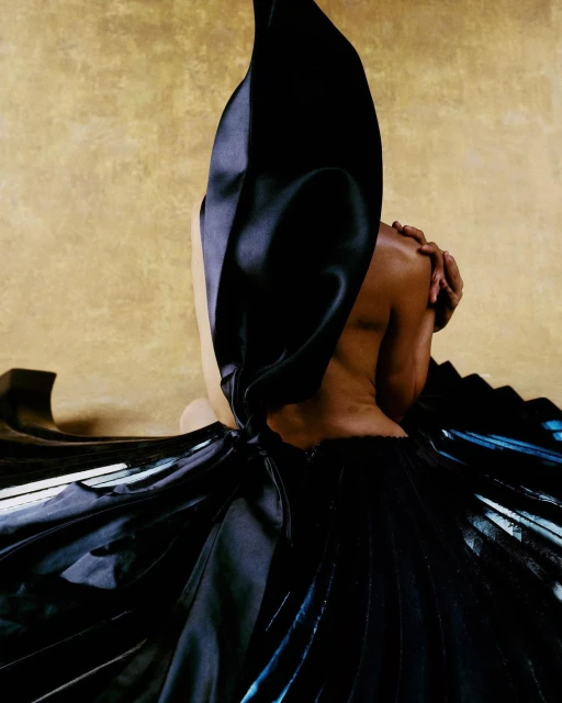 Back view of a partially visible Black woman wearing a draped black dress against a golden background in a studio setting.
