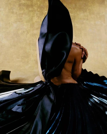 Back view of a partially visible Black woman wearing a draped black dress against a golden background in a studio setting.