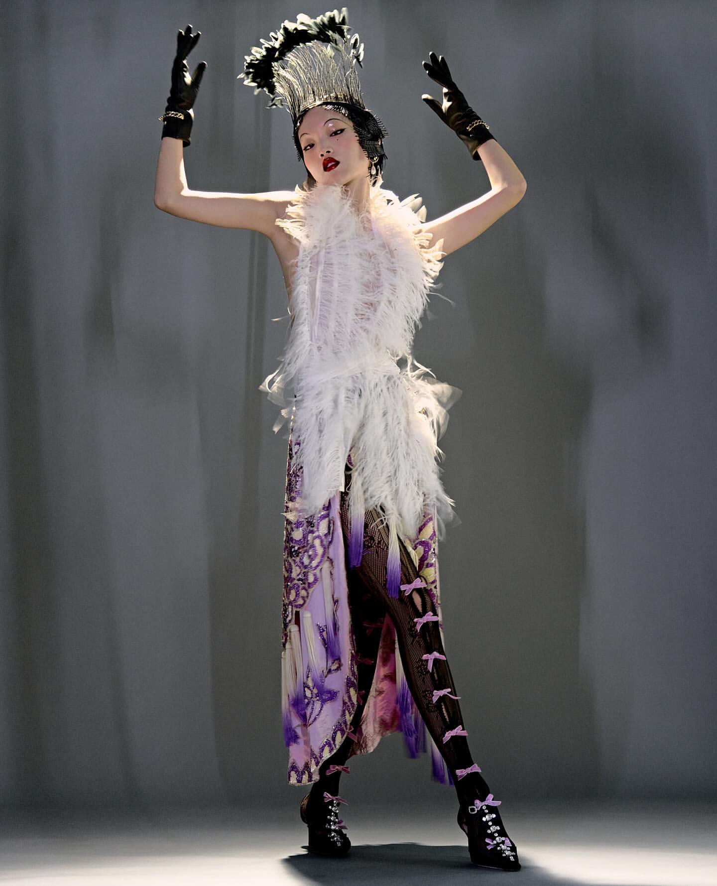 Elegant woman strikes pose in avant-garde outfit, headpiece and gloves. Feather and butterfly details, elaborate accessories, studio lighting.