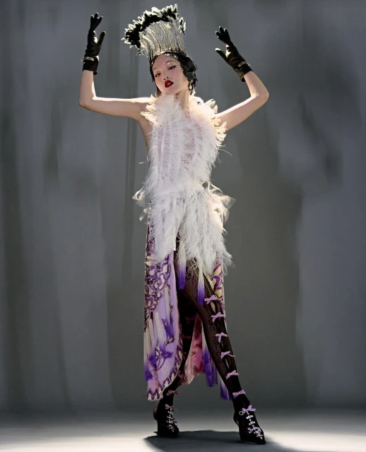 Elegant woman strikes pose in avant-garde outfit, headpiece and gloves. Feather and butterfly details, elaborate accessories, studio lighting.