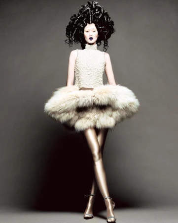 Fashion shot of a pale-skinned model in a textured top, furry skirt, tights, and heels, posing against a grey backdrop with dramatic makeup.