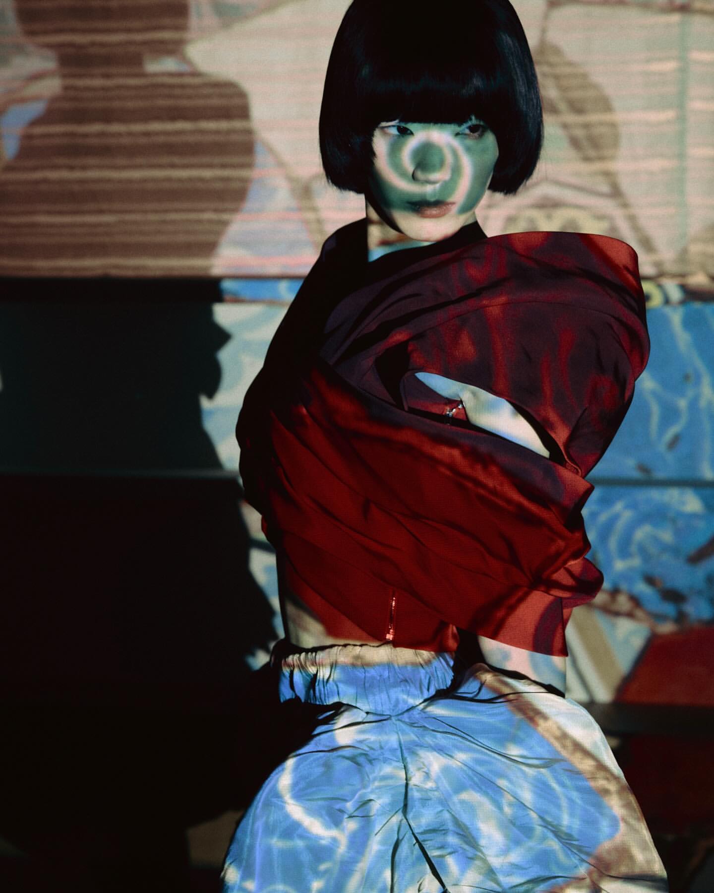 Artistic fashion portrait of a model with face paint, wearing a red shawl and blue skirt, against a patterned backdrop.