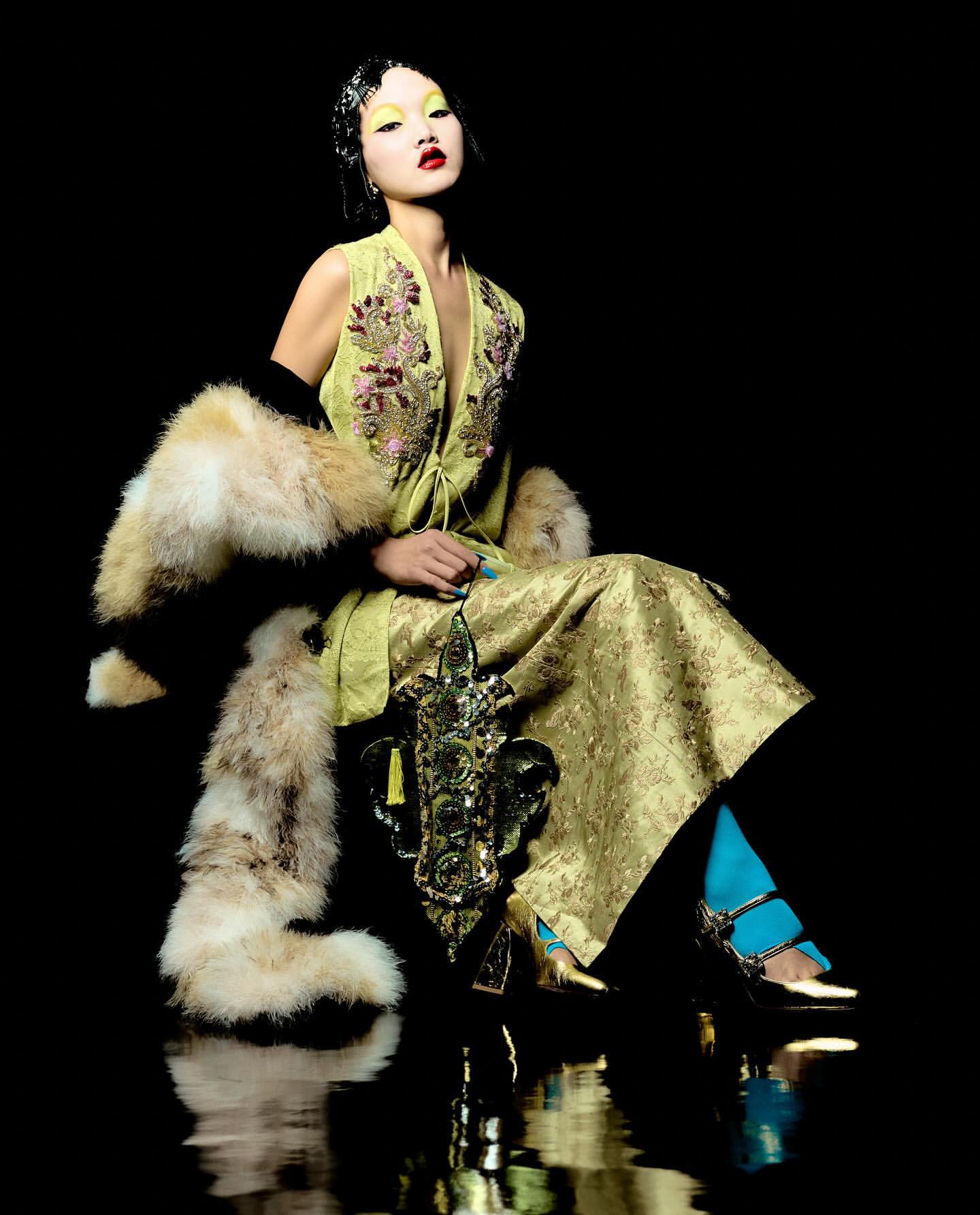Asian woman in embroidered green dress and fur stole sits in studio with ornate prop, dramatic lighting, reflective floor, fashion shot.