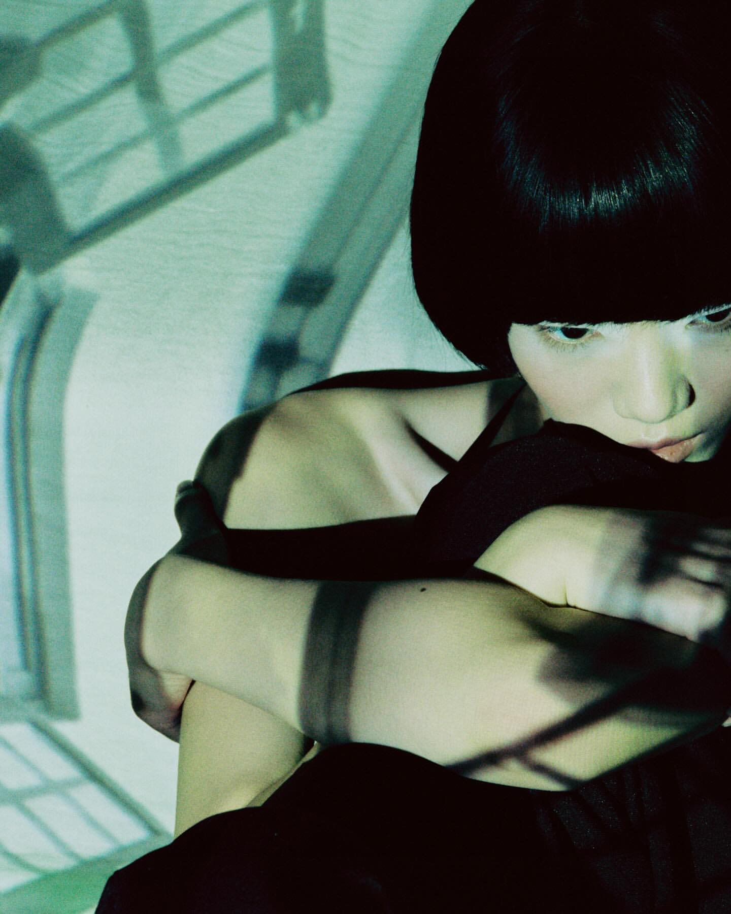 Portrait of a woman with short dark hair, hugging her knees in a darkened room with window shadows, creating a moody atmosphere.
