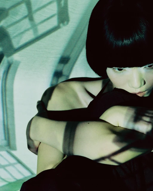 Portrait of a woman with short dark hair, hugging her knees in a darkened room with window shadows, creating a moody atmosphere.