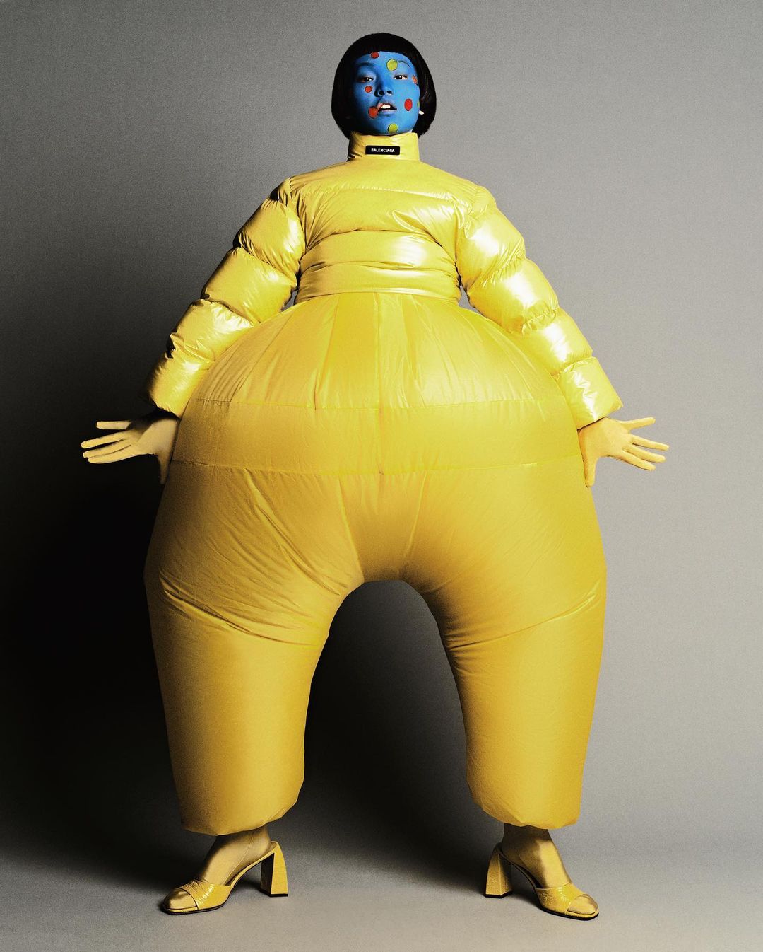 Model with blue face paint poses in an oversized yellow puffer suit and yellow heels against a grey background in a studio fashion shot.