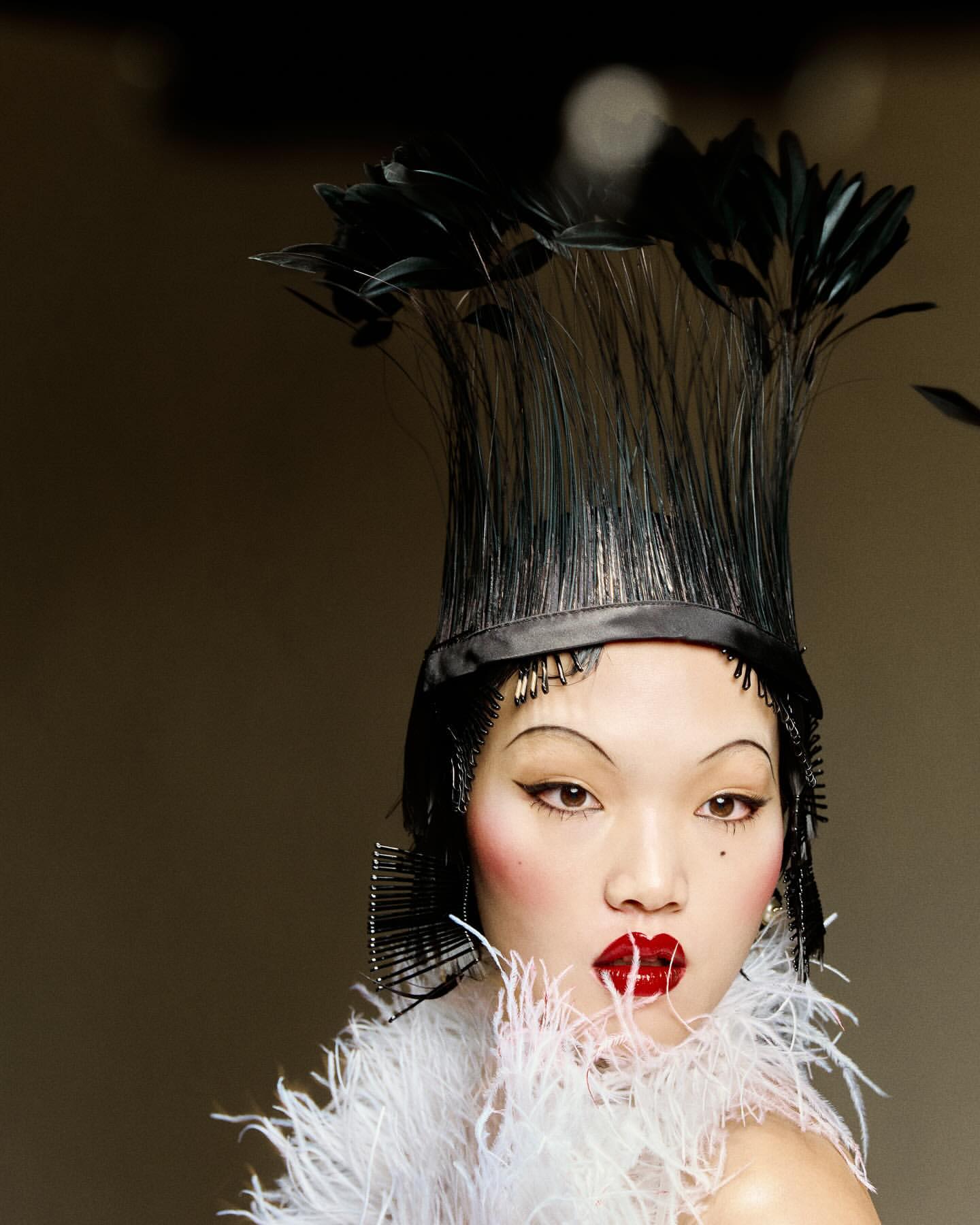 Asian woman with bright red lipstick, a dramatic feather collar, and a tall feathered hat poses in front of a neutral grey background.