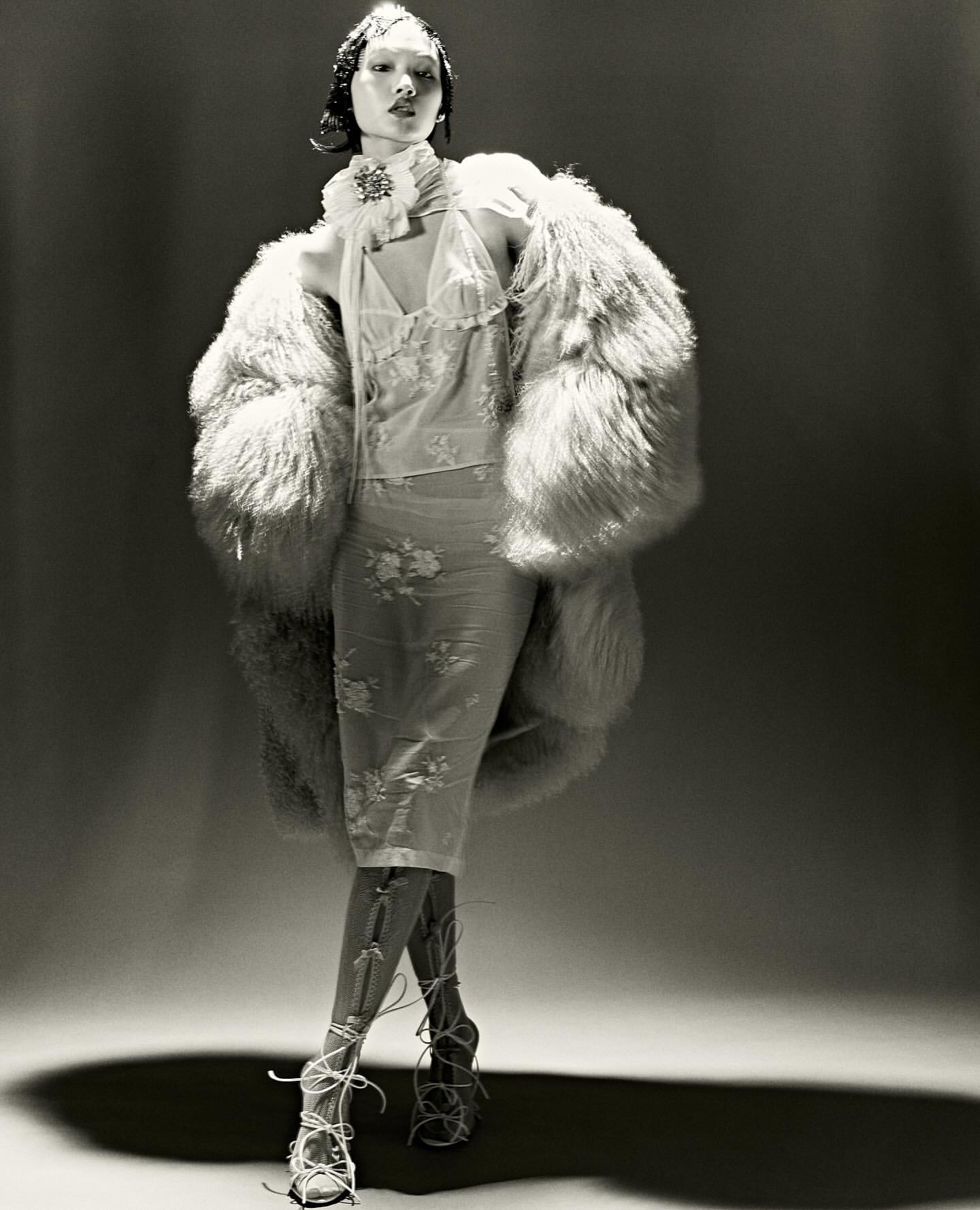 A model stands in a photo studio wearing a sheer dress, a large fur coat, and lace-up heels, captured in a monochrome studio shot.