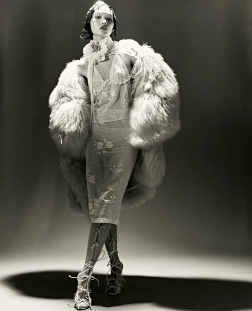 A model stands in a photo studio wearing a sheer dress, a large fur coat, and lace-up heels, captured in a monochrome studio shot.