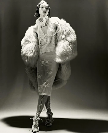 A model stands in a photo studio wearing a sheer dress, a large fur coat, and lace-up heels, captured in a monochrome studio shot.