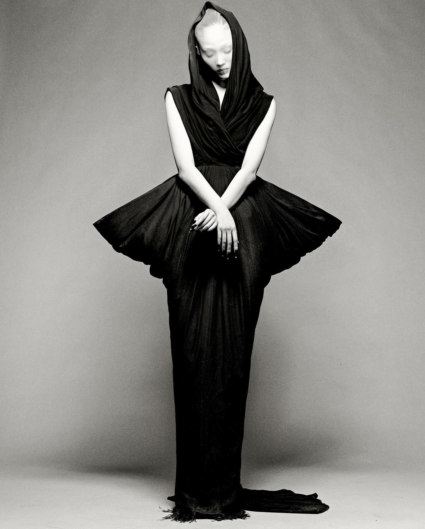 Monochrome fashion shot of a slender adult woman in a dramatic hooded black gown with a wide, pleated skirt and train on grey backdrop.