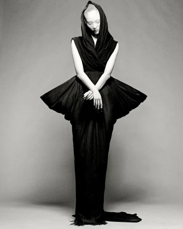Monochrome fashion shot of a slender adult woman in a dramatic hooded black gown with a wide, pleated skirt and train on grey backdrop.