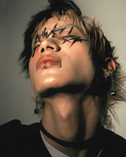 Close-up portrait of a person with creative black graphic eye makeup, lip and ear piercings, and a black choker in a studio setting.