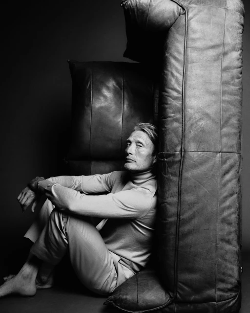 Monochrome portrait of Mads Mikkelsen seated on the floor with his arms crossed next to a stack of leather cushions in studio lighting.