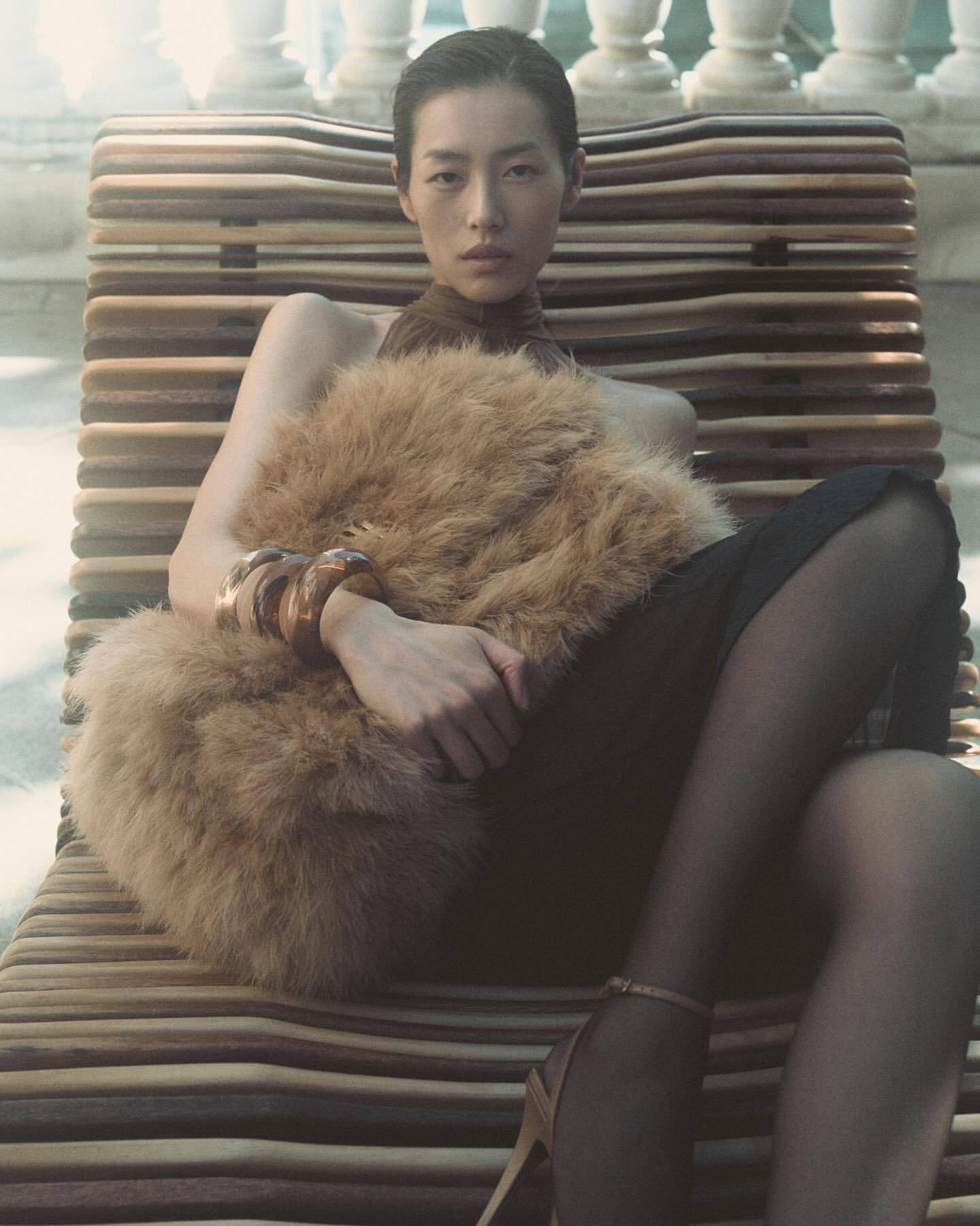 Asian woman poses in a brown faux fur stole, black dress, and high heels, resting on a wooden lounger with balustrade background.