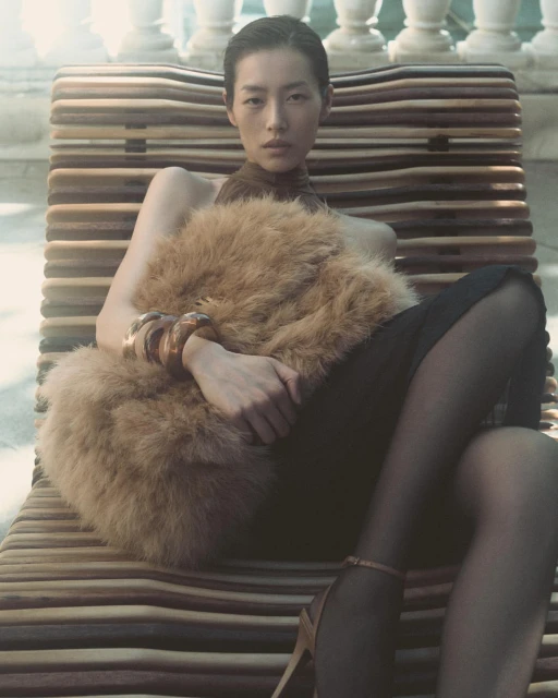 Asian woman poses in a brown faux fur stole, black dress, and high heels, resting on a wooden lounger with balustrade background.