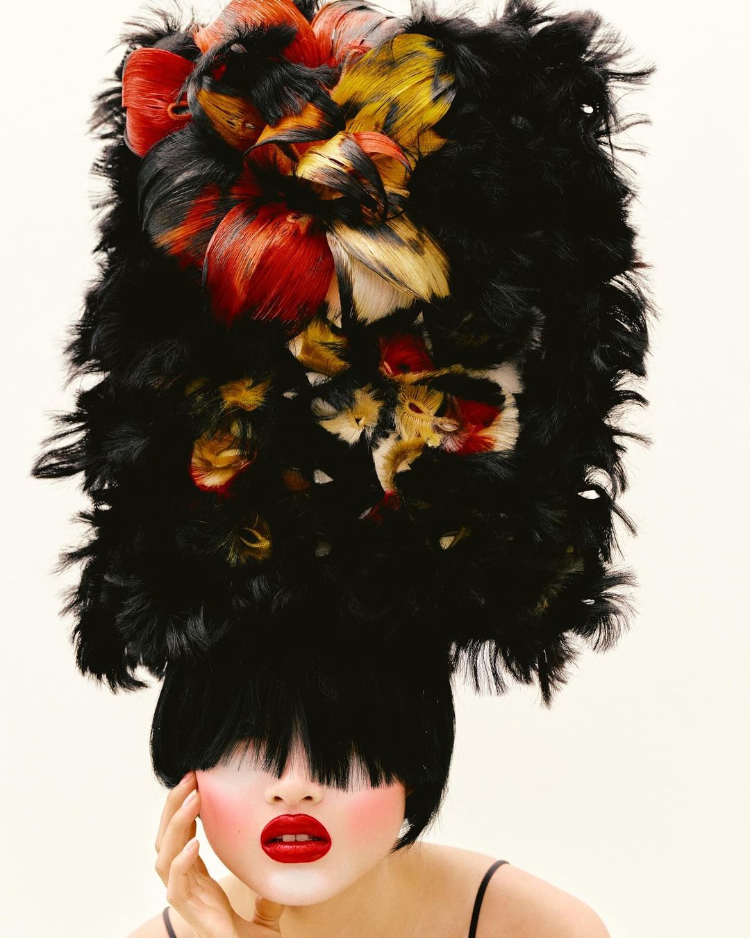 Portrait of a woman with dark hair, red lipstick, and pale skin, wearing a large, colorful feathered headpiece against a white background.