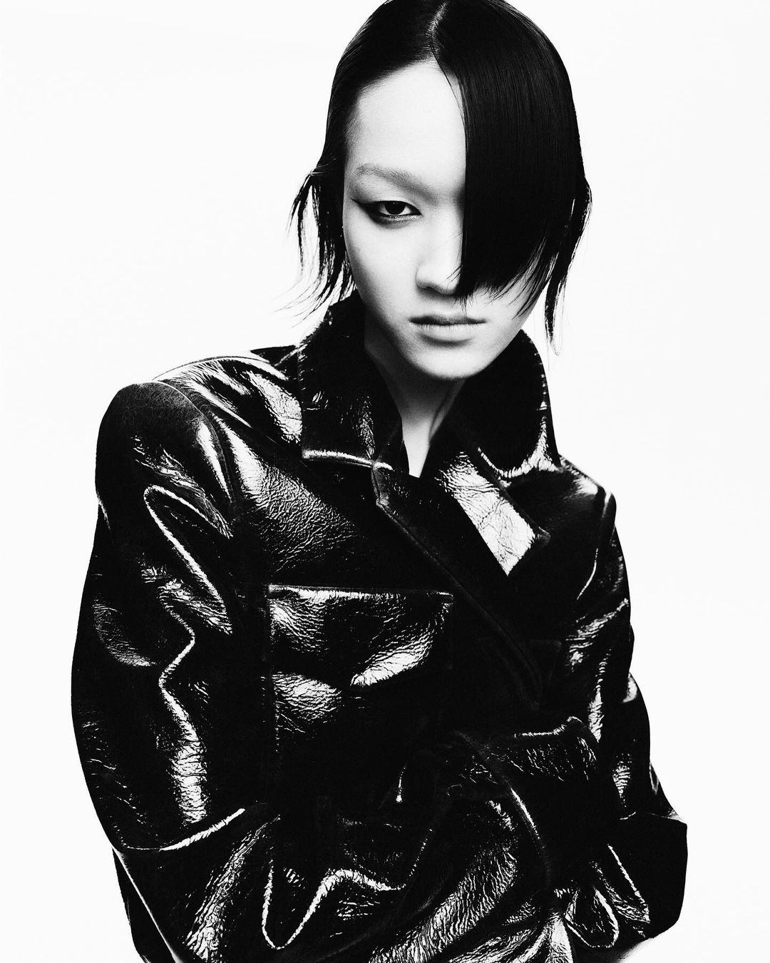 Portrait of an Asian woman with dark hair, wearing a shiny black trench coat against a white background in a studio fashion shot.