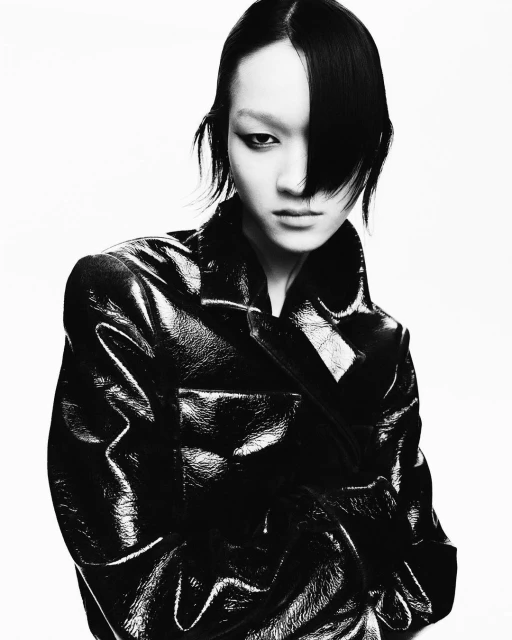 Portrait of an Asian woman with dark hair, wearing a shiny black trench coat against a white background in a studio fashion shot.