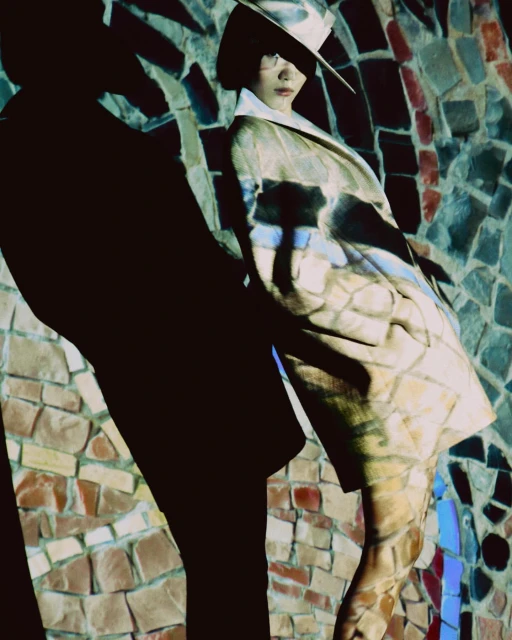Woman in beige hat and coat poses against mosaic wall with projected shadow, fashion photography, studio lighting, and geometric patterns.