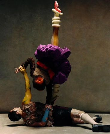 Two figures performing in a studio setting; one lies on the floor supporting the other, who balances atop a custom headpiece.