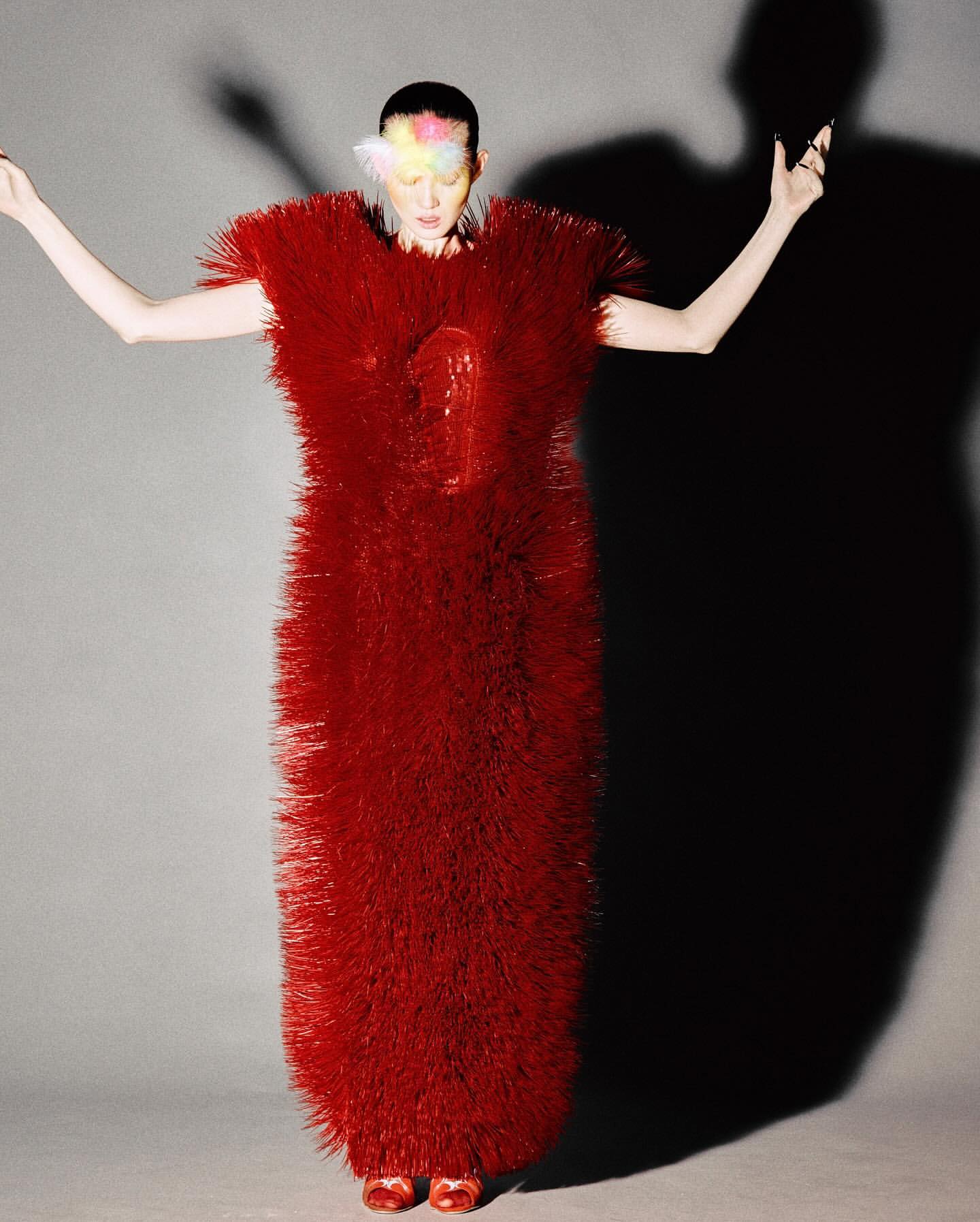 Woman in an avant-garde red dress stands in a studio with arms raised, a colorful headpiece, and a dark shadow behind her.