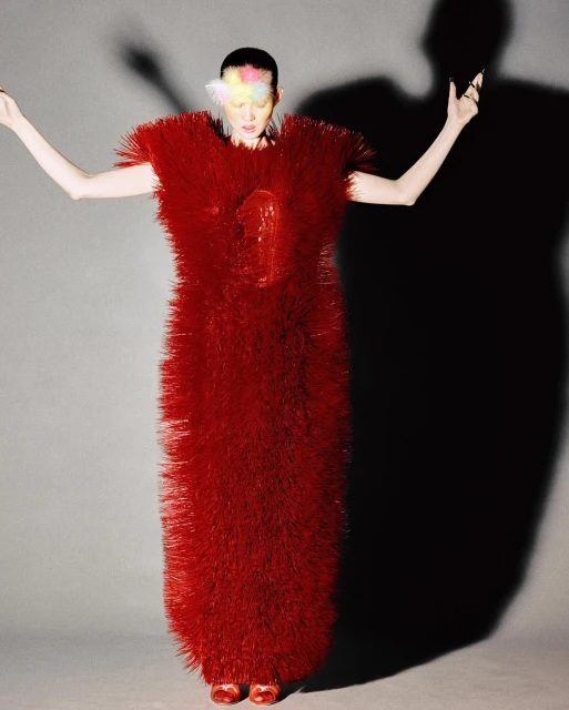 Woman in an avant-garde red dress stands in a studio with arms raised, a colorful headpiece, and a dark shadow behind her.