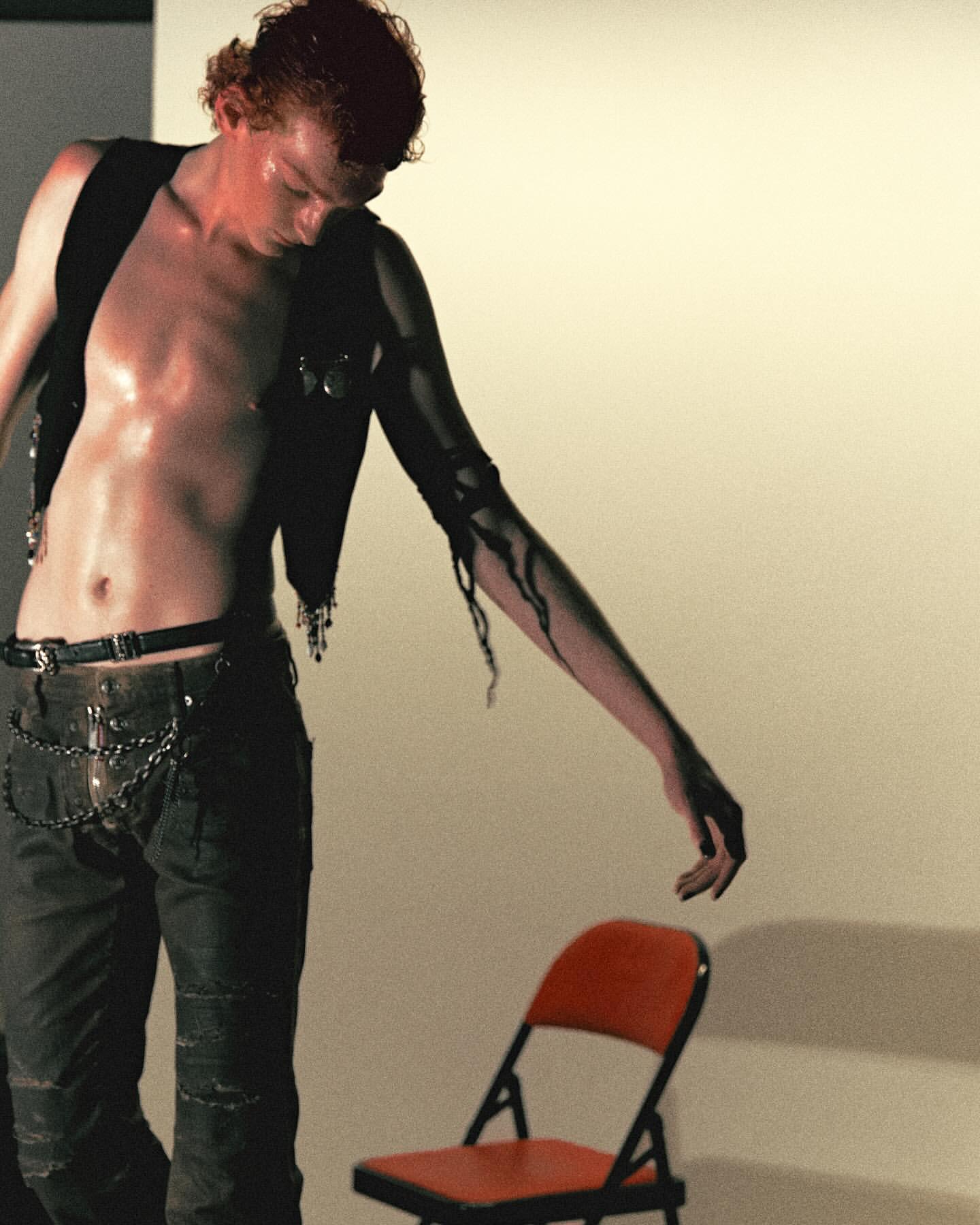 An adult male with black paint on his arm and wearing a black vest poses next to a red chair against a neutral backdrop in a studio setting.