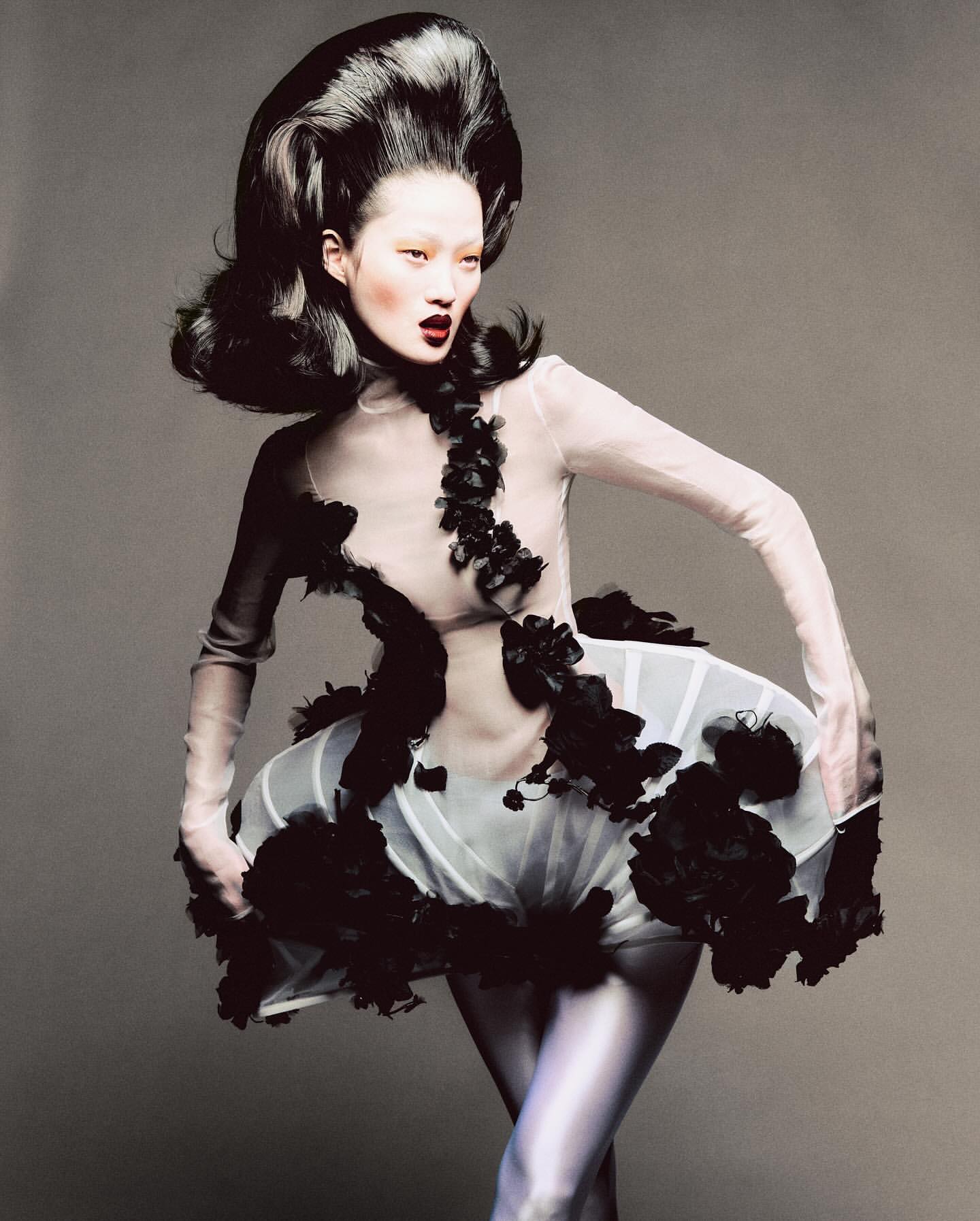 Avant-garde fashion portrait of an Asian woman with voluminous black hair, wearing a sheer white dress with black floral embellishments, studio shot.