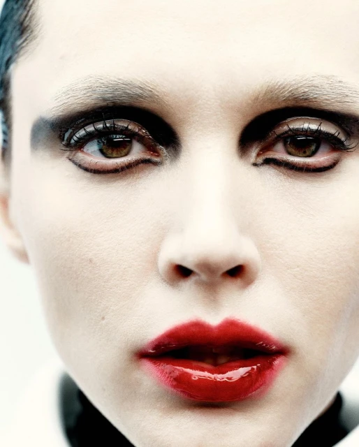 Close-up portrait of a woman with fair skin, dramatic black eye makeup, and glossy red lips against a bright white background.