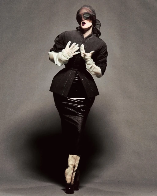 A fashionably dressed person in a black jacket and shiny skirt poses against a grey backdrop, face obscured by large dark glasses.