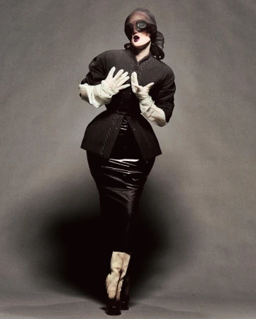 A fashionably dressed person in a black jacket and shiny skirt poses against a grey backdrop, face obscured by large dark glasses.