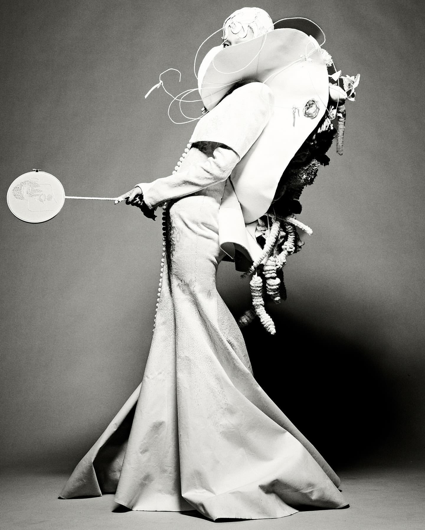 Monochrome fashion portrait of a person in an extravagant white dress with sculptural details, holding a small, round fan against a grey backdrop.
