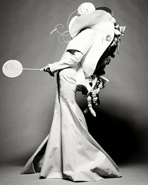Monochrome fashion portrait of a person in an extravagant white dress with sculptural details, holding a small, round fan against a grey backdrop.