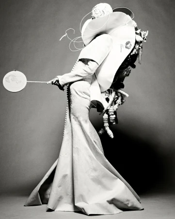 Monochrome fashion portrait of a person in an extravagant white dress with sculptural details, holding a small, round fan against a grey backdrop.