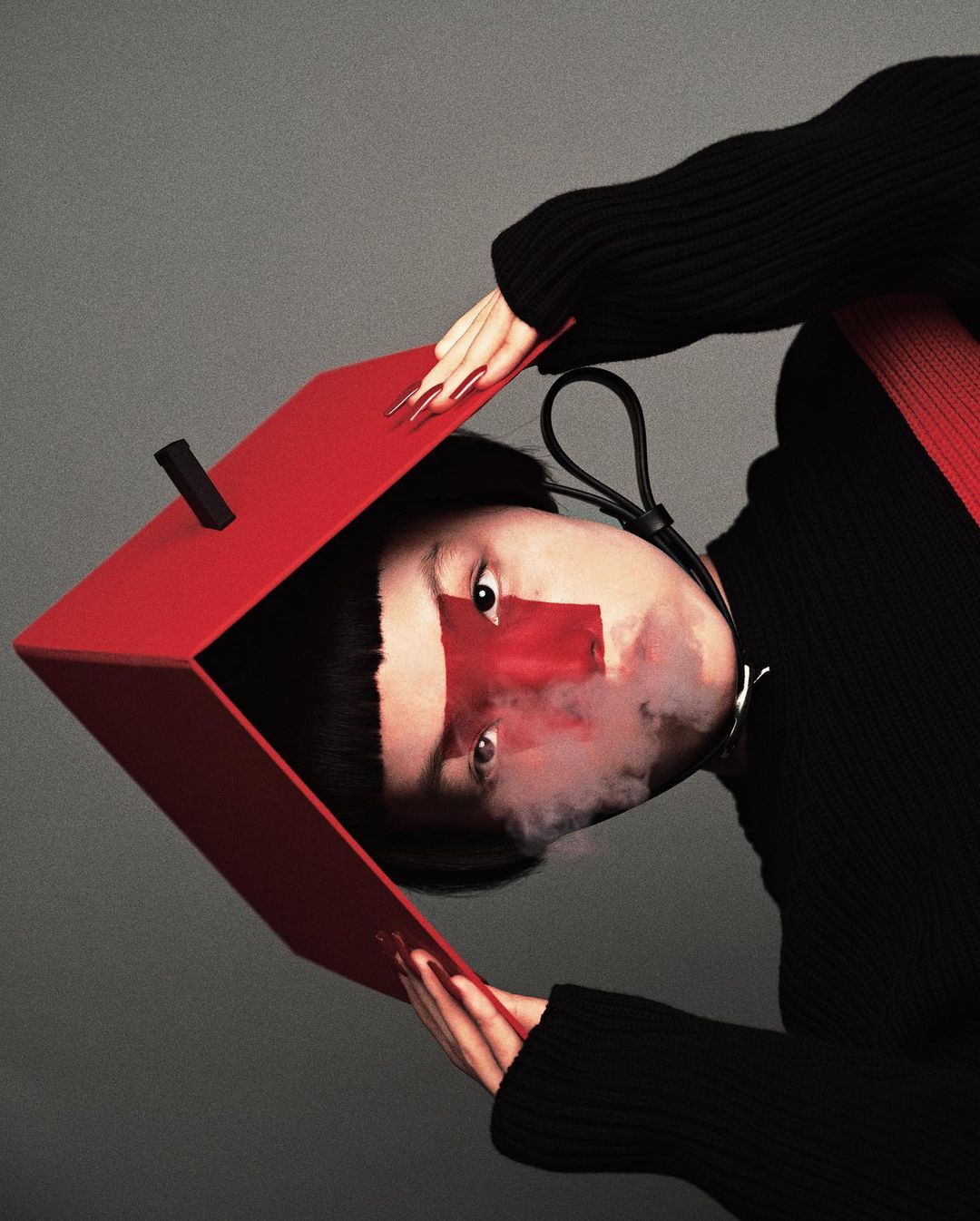 Woman with short black hair posing in a black long-sleeved shirt, a red square over her eye, holding a large red cubic box.